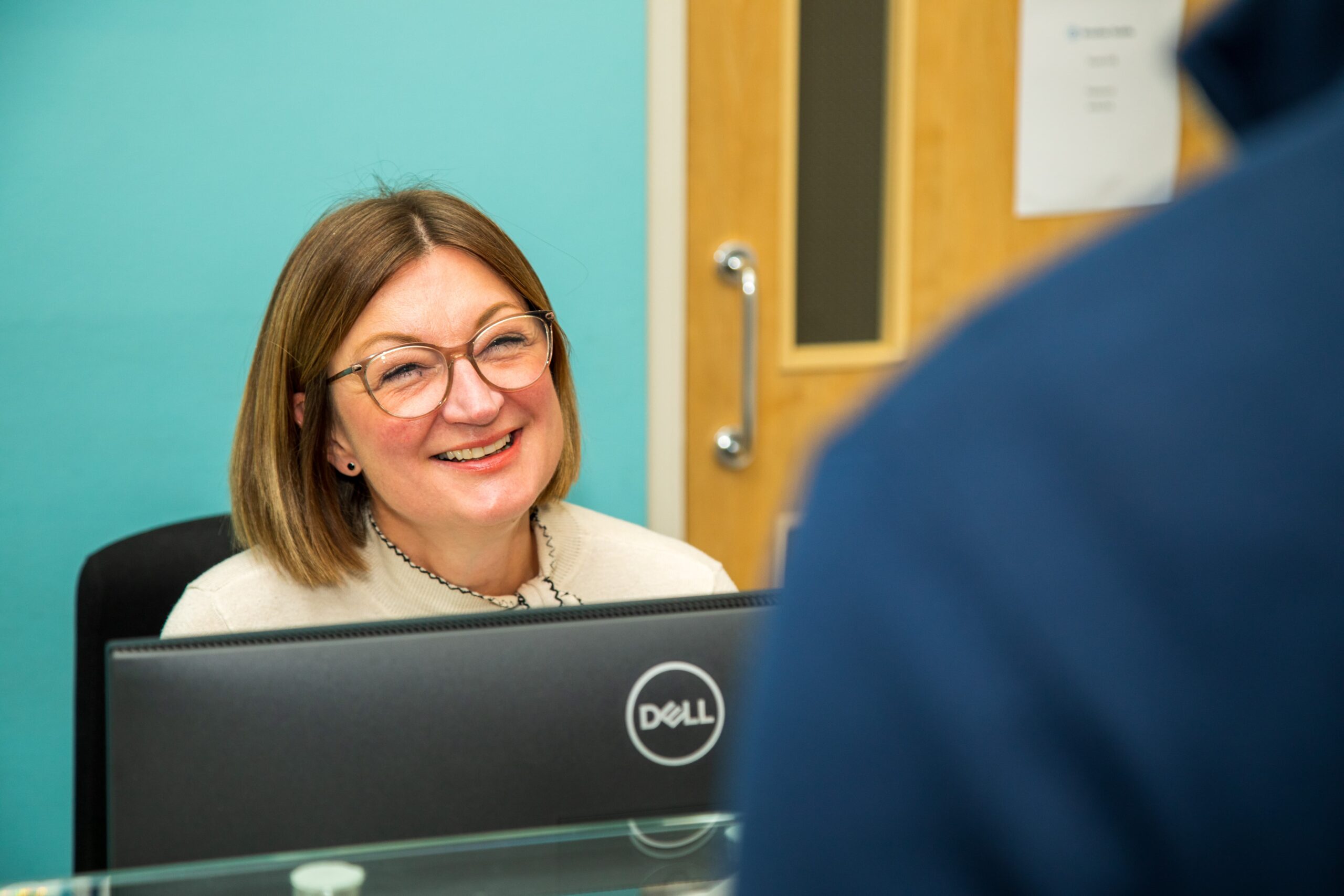 A receptionist sits behind a desk with a computer monitor, speaking with someone standing on the other side in an office setting.