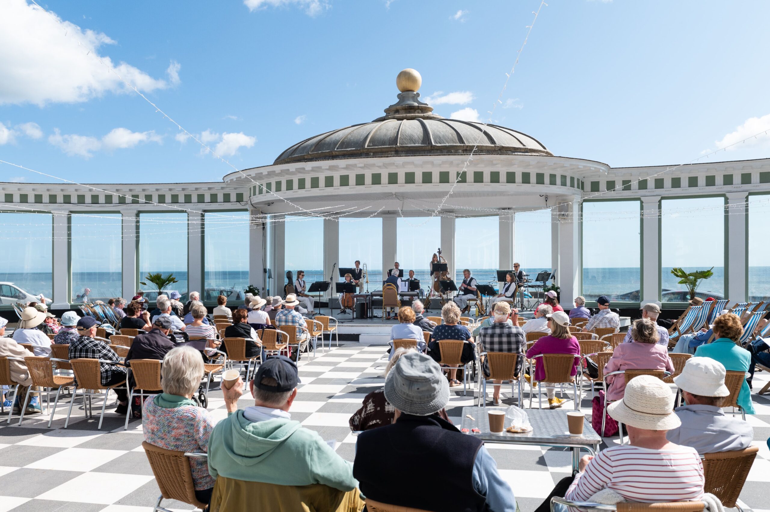 An audience sits on a sunlit terrace overlooking the sea, watching a live performance by a small orchestra beneath a domed open‑air pavilion