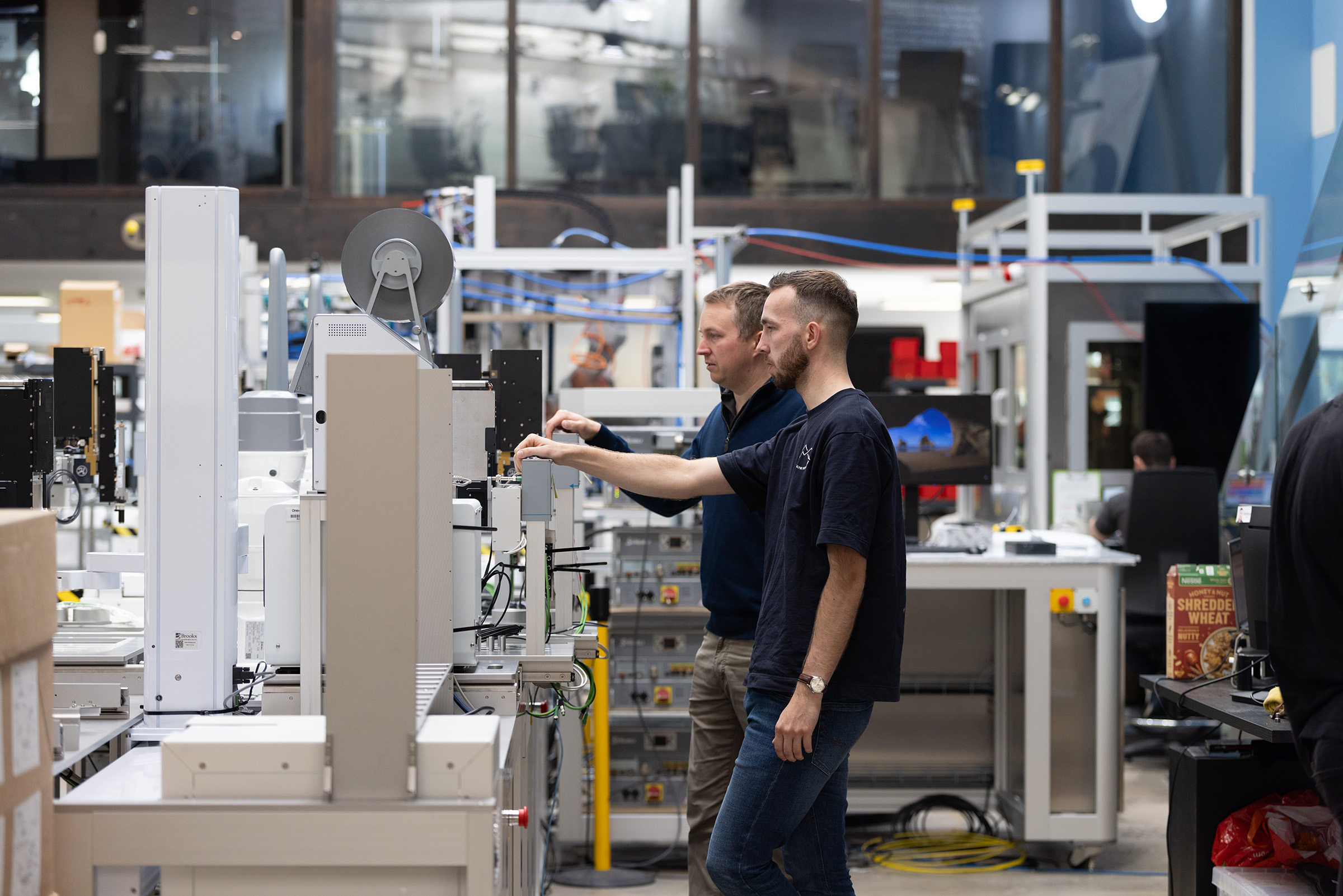 Two people stand at a workstation in a modern industrial facility, adjusting equipment surrounded by machinery and electronic components.