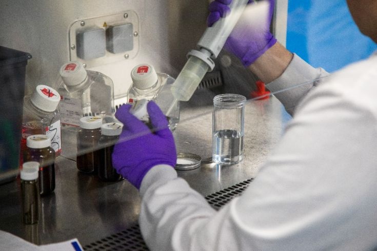 A person wearing purple gloves works at a laboratory station, using a pipette to transfer liquid into a container surrounded by bottles and equipment. Image credit: Labcorp