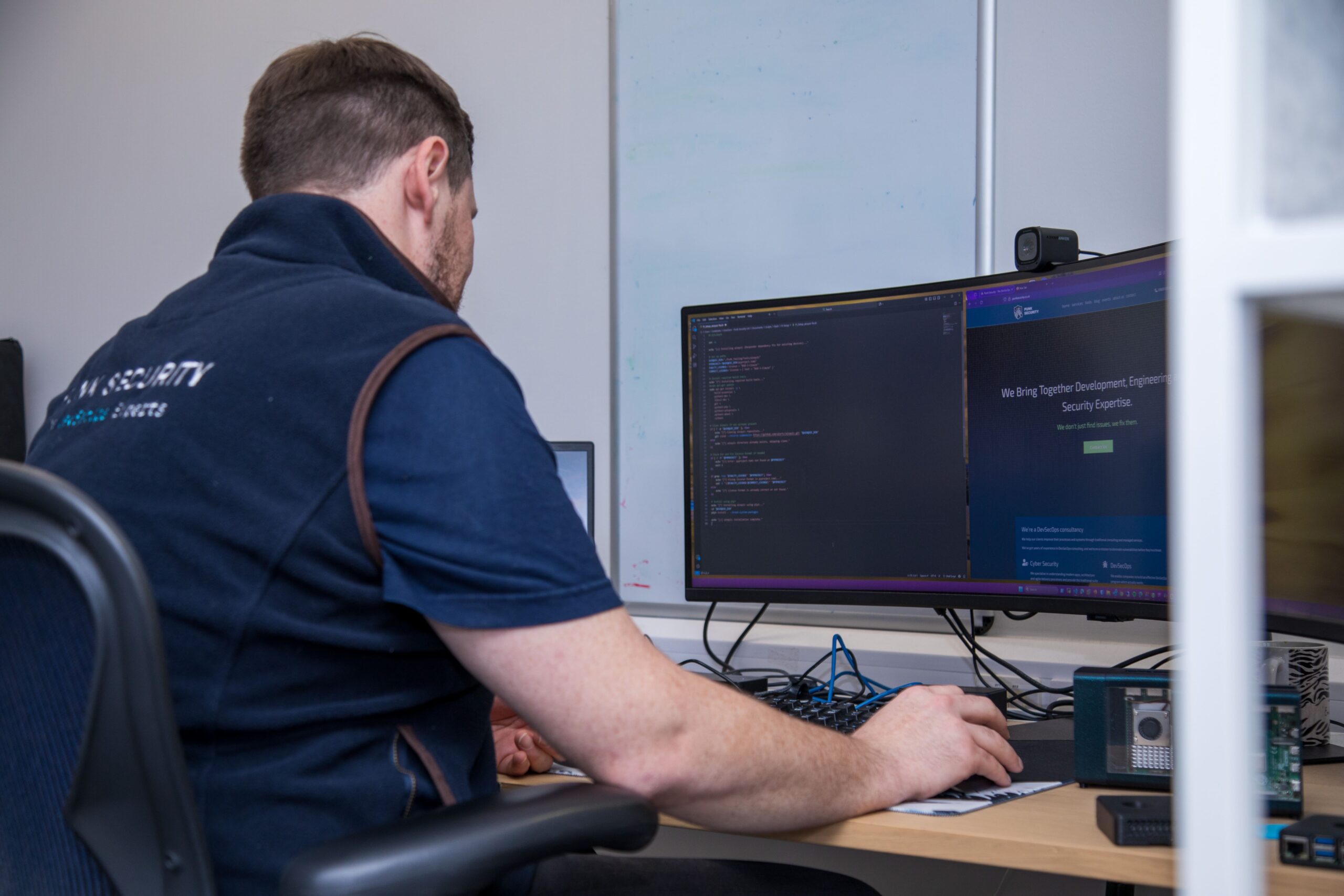 A person sits at a dual‑monitor workstation, working on code and cybersecurity tools in an office setting.