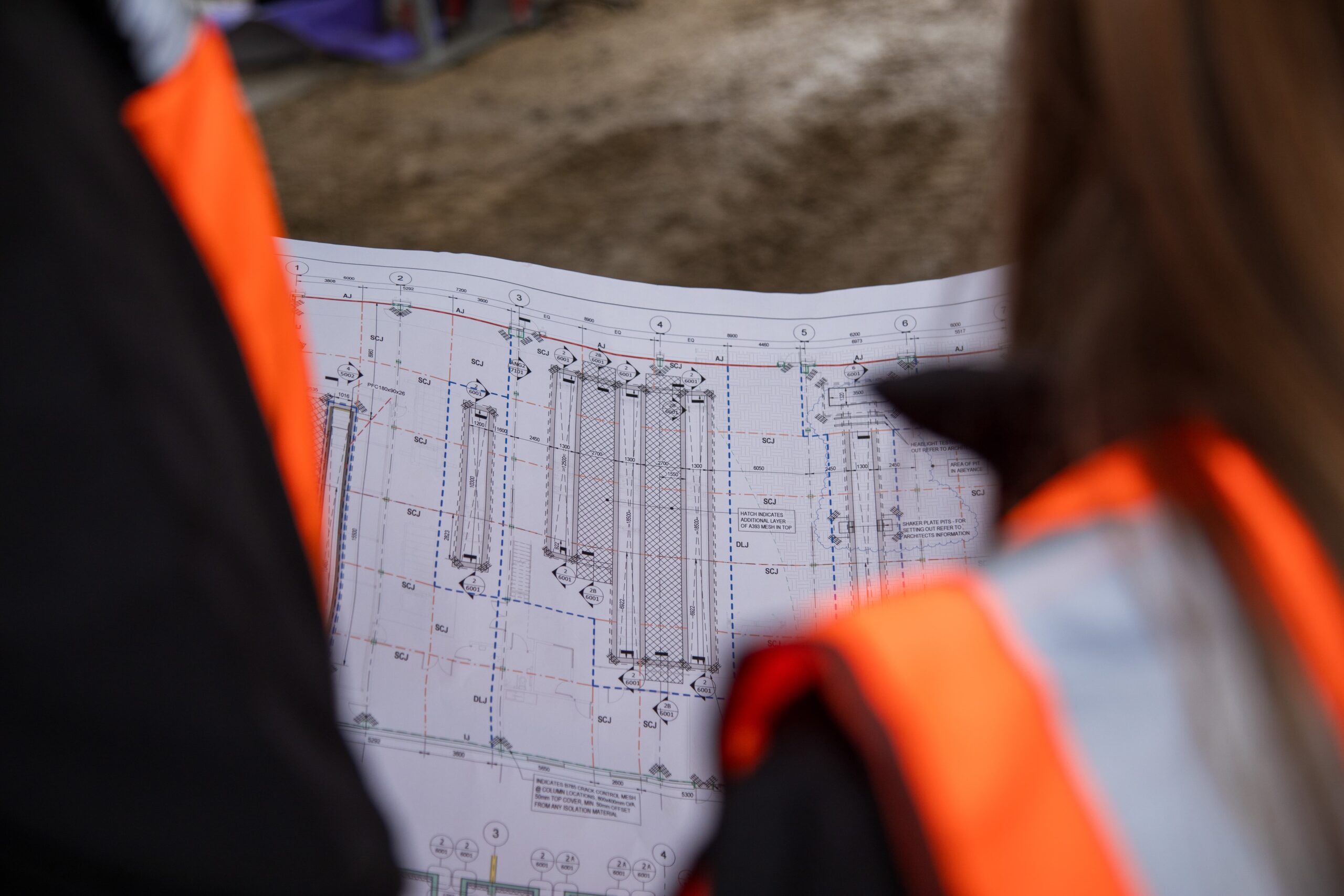 Two people in high‑visibility vests examine a detailed construction blueprint showing structural drawings and measurements at a site. Image credit: NYC and HACS Group