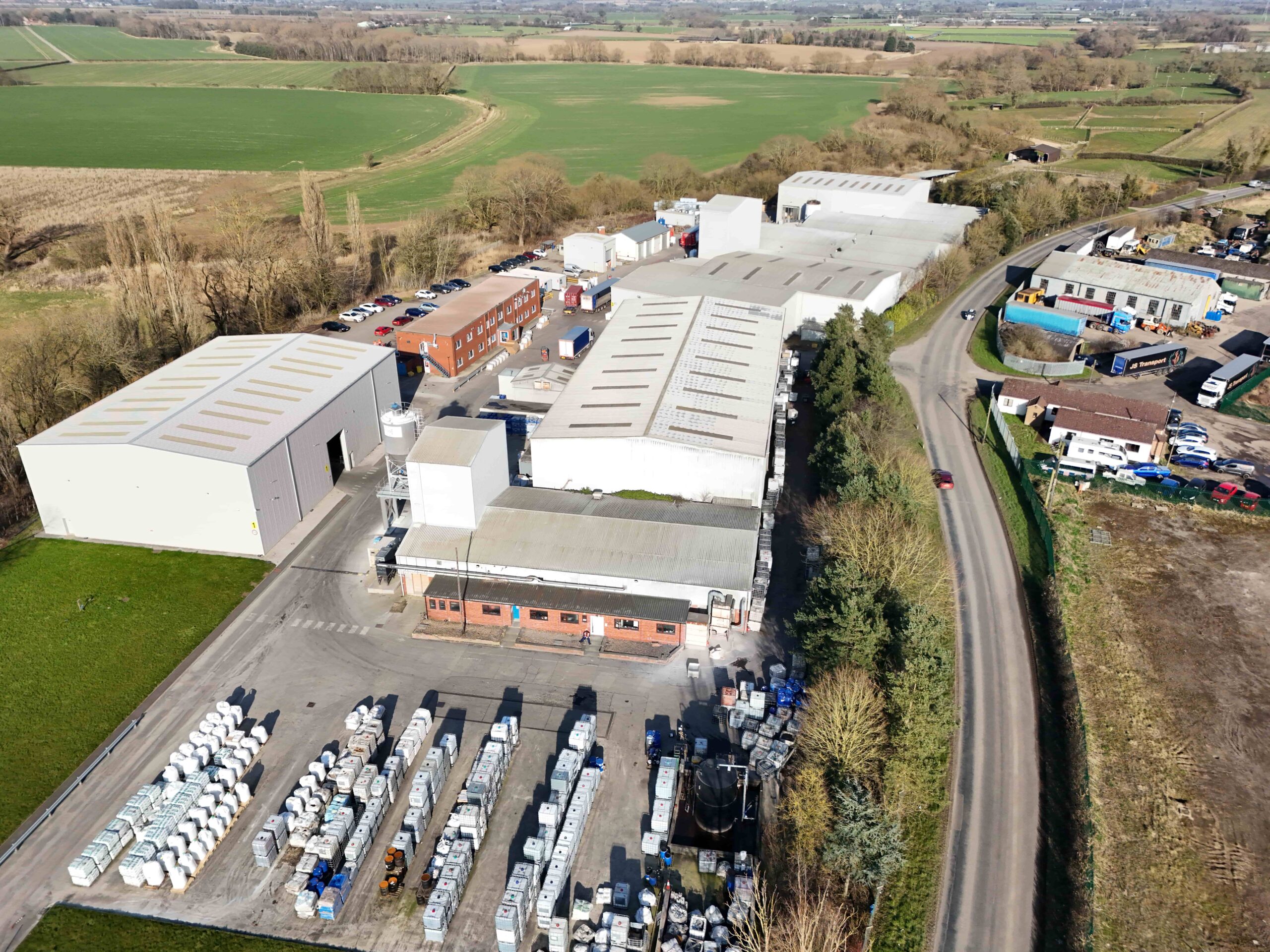Aerial view of an industrial site with several large warehouse buildings, outdoor storage areas, a car park, and surrounding open fields along a rural road.