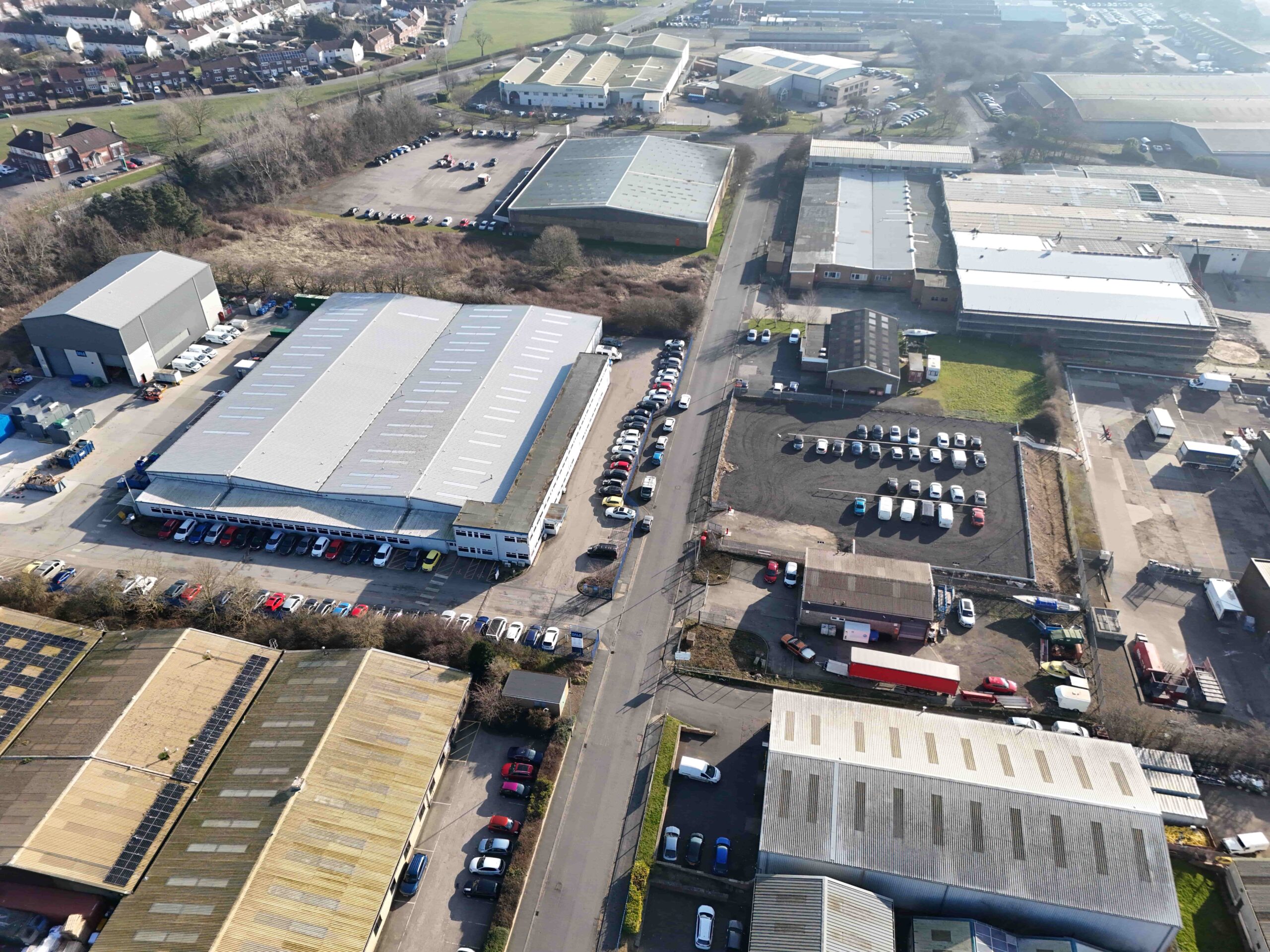 Aerial view of an industrial estate with several warehouse buildings, parking areas, and nearby residential areas in the background