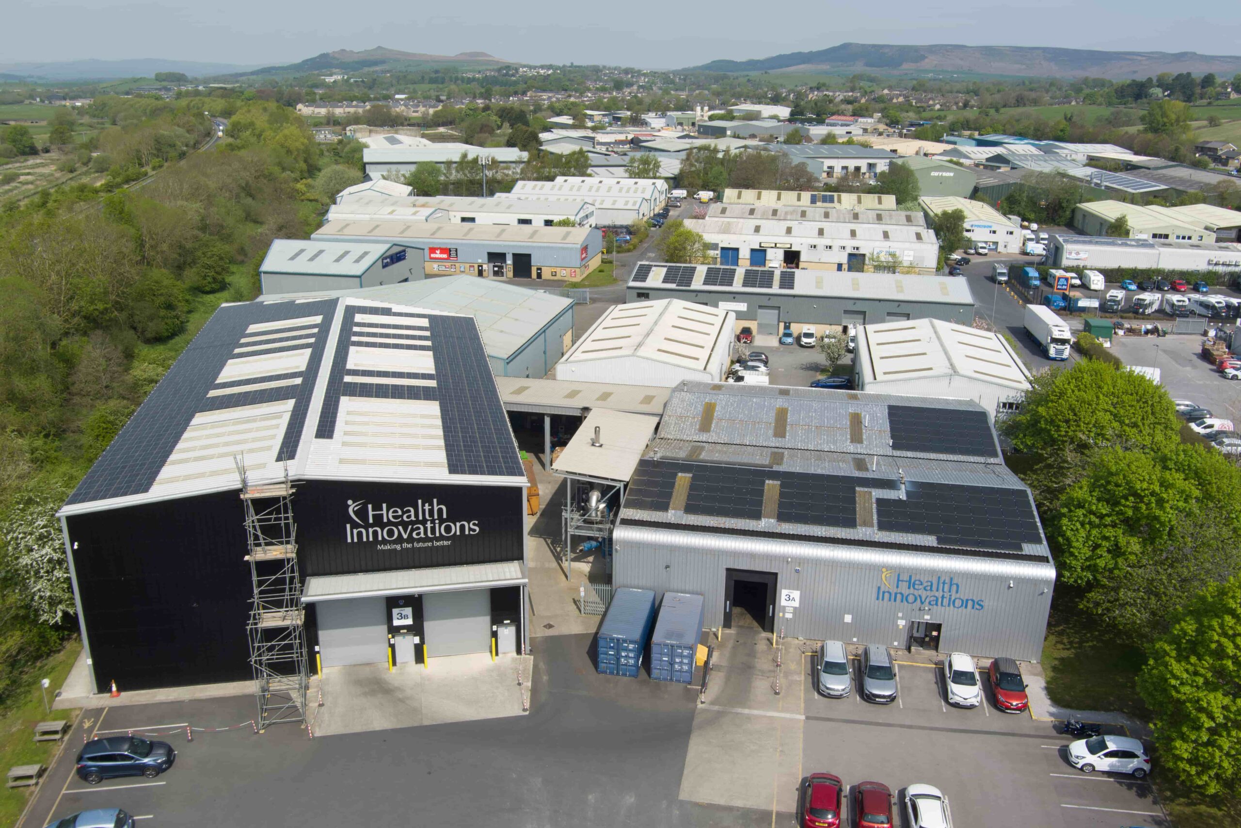 Aerial view of an industrial estate with multiple warehouse-style buildings surrounded by trees and distant hills. Two prominent buildings in the foreground display ‘Health Innovations’ signage. Many rooftops are fitted with solar panels. Several parked vehicles and shipping containers are visible around the site.