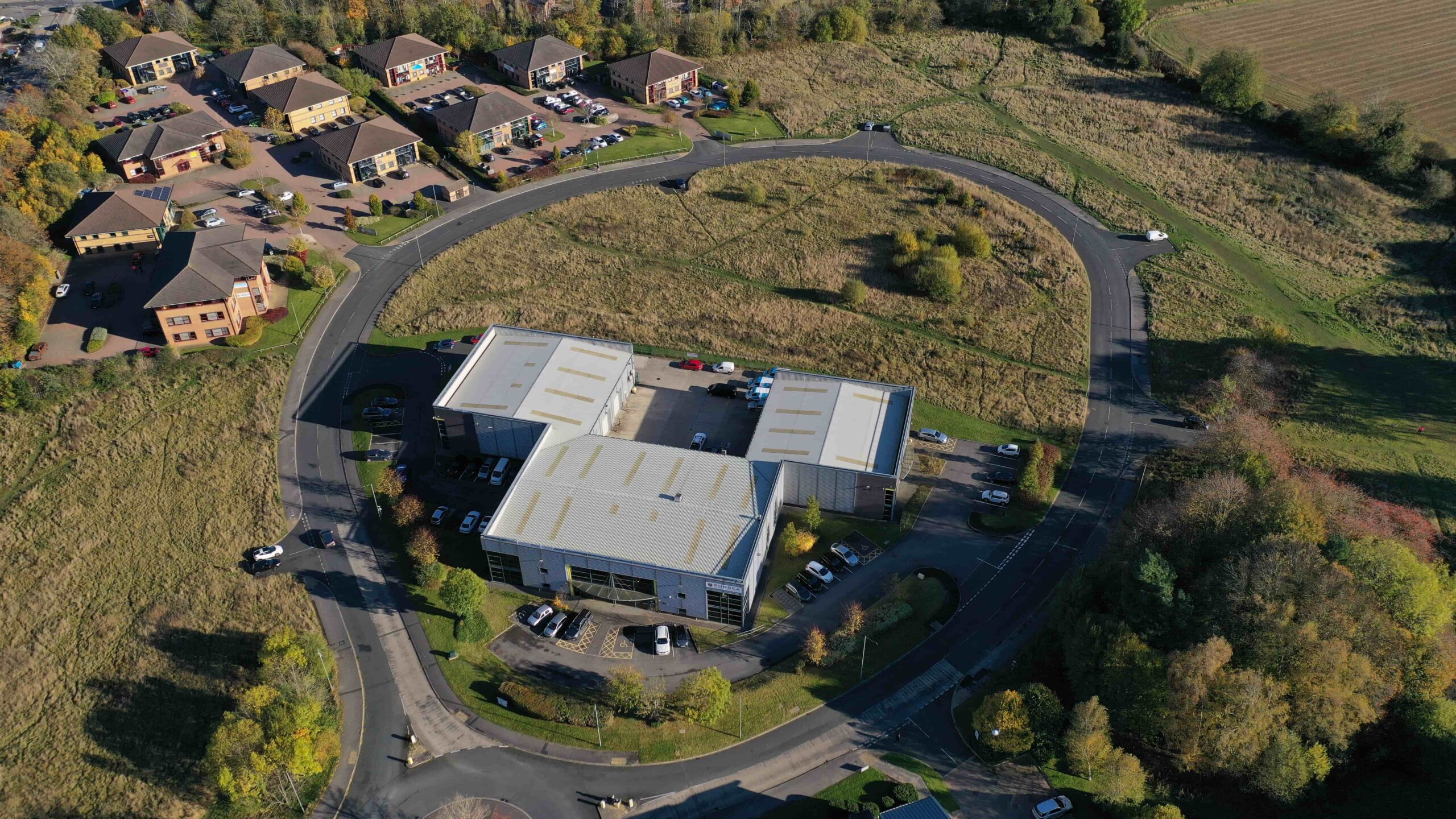 Aerial photograph showing a large industrial unit in a central green space with 2 and 4 story office blocks located in the northwest of the site and a business centre located in the southeast of the site.