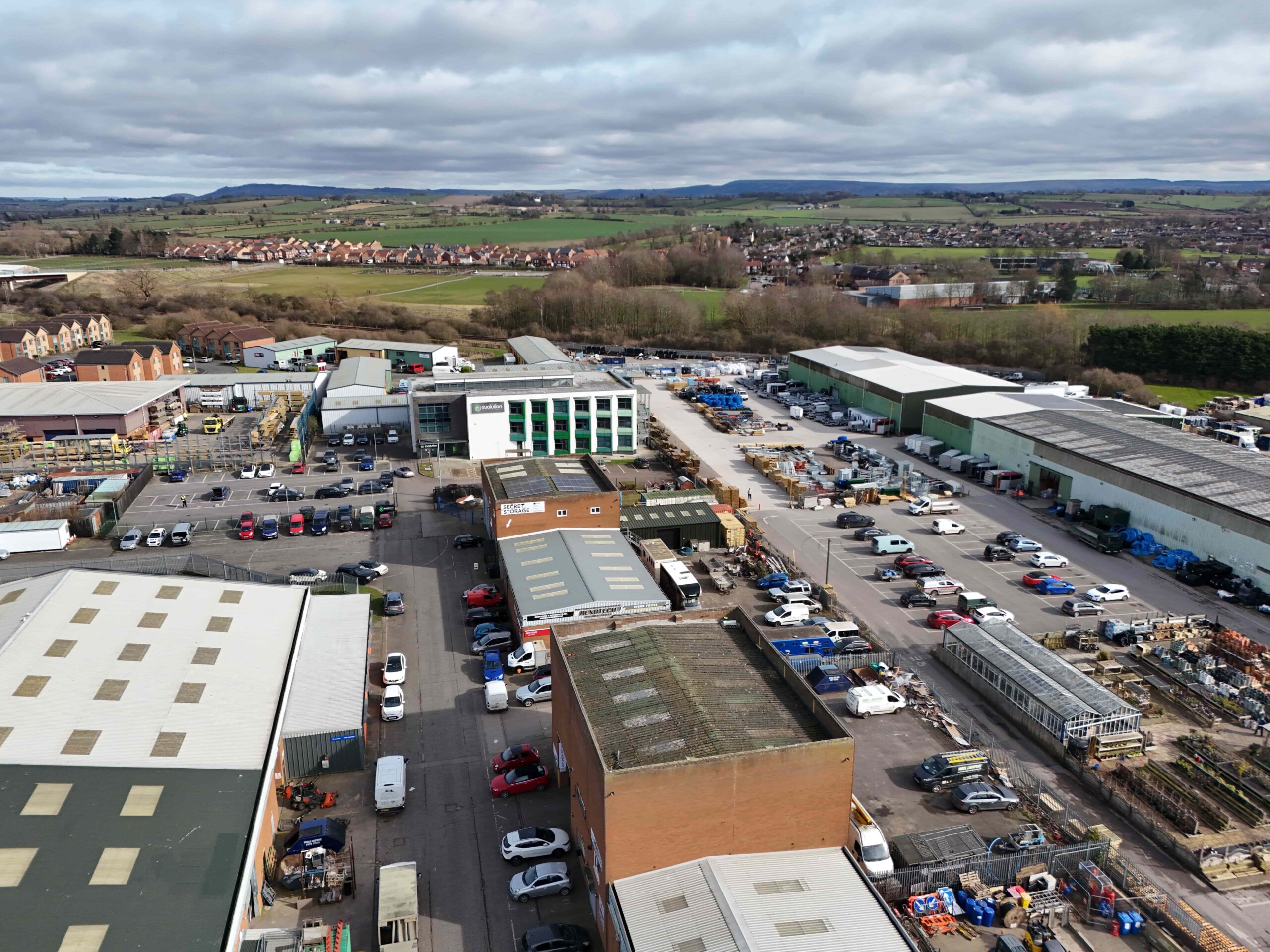 Aerial view of an industrial estate with multiple warehouse buildings, parked vehicles, and surrounding fields and housing in the distance.