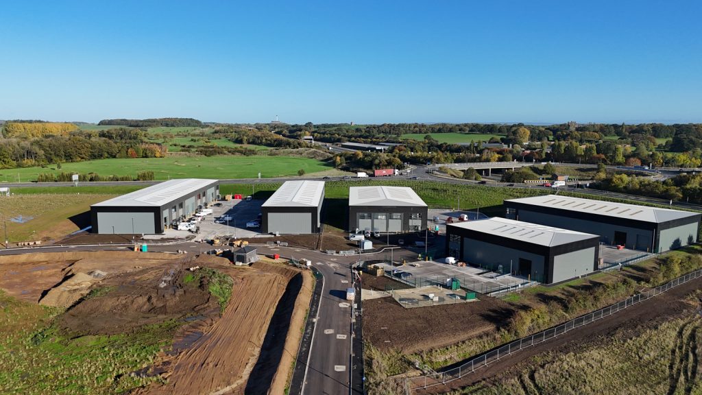 Aerial view of a construction site featuring several newly built industrial-style units with grey roofs, arranged around newly laid roads. Surrounding the site are open green fields and distant trees under a clear blue sky.