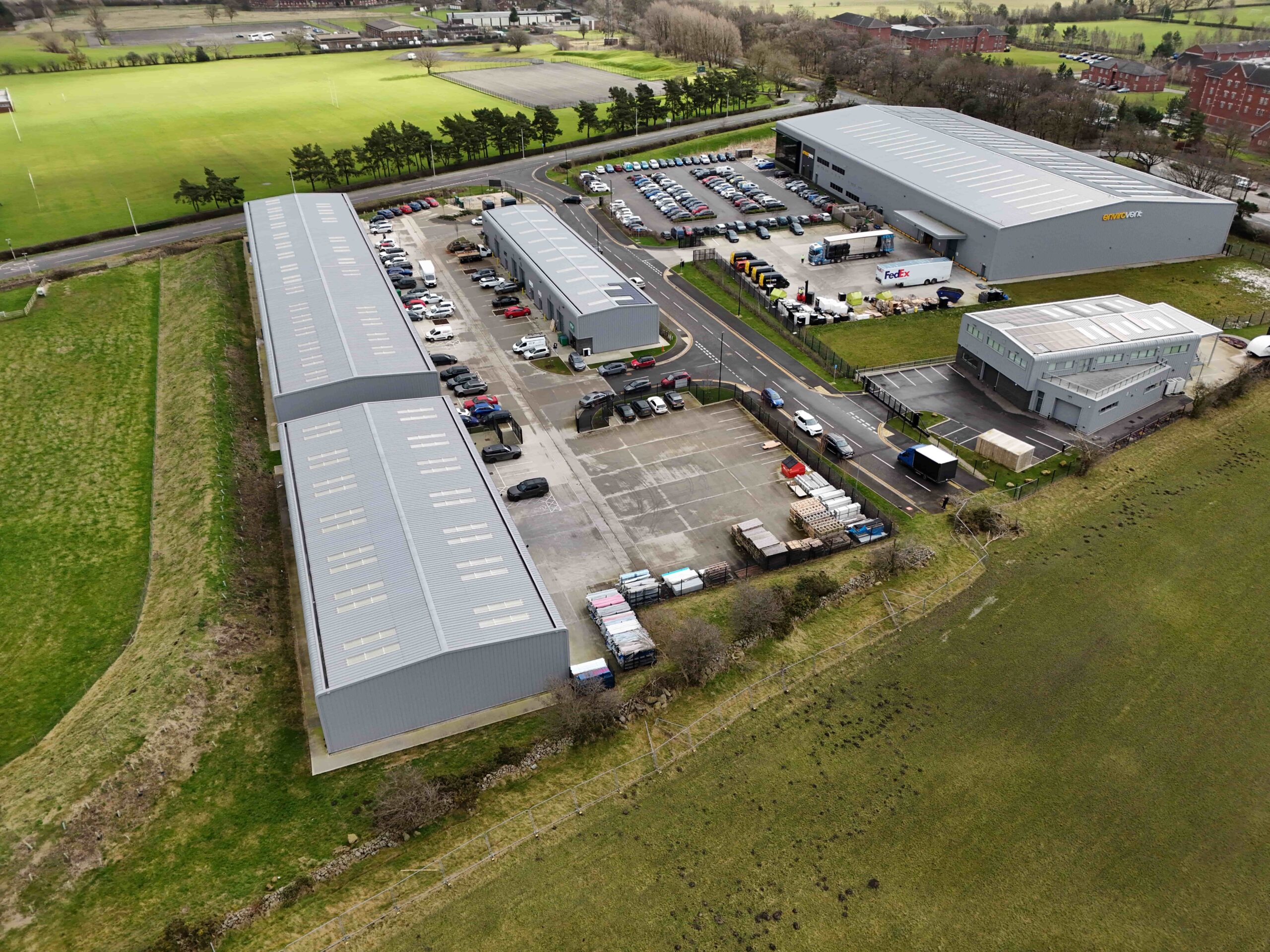 Aerial view of an industrial business park with several large grey warehouse buildings, parking areas, and surrounding green fields.